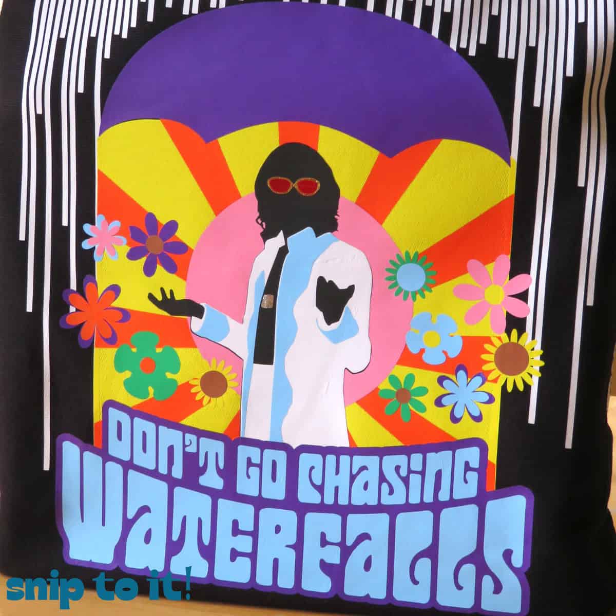 closeup of an abstract design made up of layered HTV on a black canvas tote bag, featuring a figure in a white suitearing sunglasses surrounded by flowers, captioned Don't go chasing waterfalls