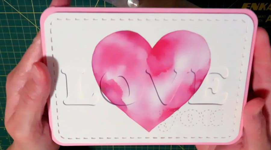 photo of completed recessed text card, LOVE in Cooper Black font cut out of an image of a pink watercolor heart with a white background. cutouts that look like stitches are around the border and also spell out "you" in Cooper Black under the LOVE.