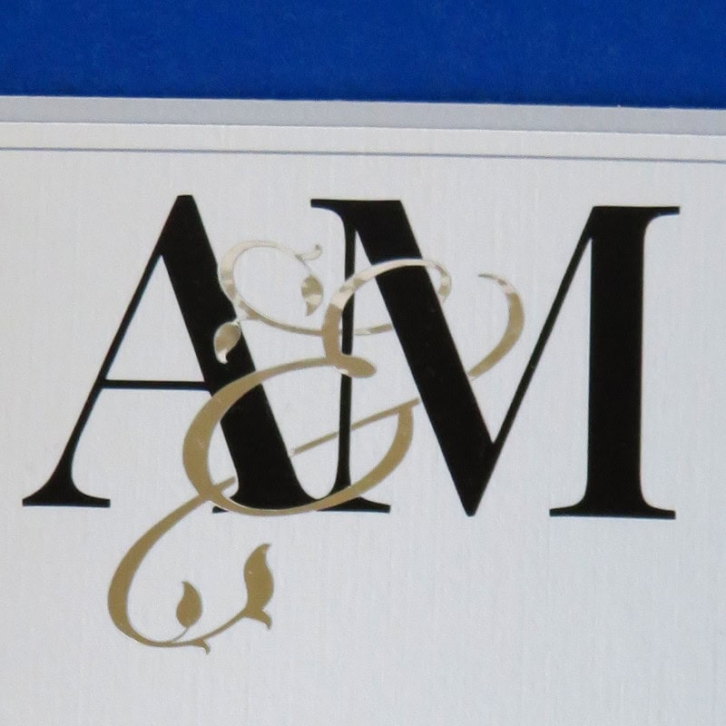 closeup image of a finished interwoven couples monogram applied to wedding invitation. The ampersand character, cut out of silver foil vinyl in a flowing script font, appears to encircle the capital letters AM, cut out of black vinyl in a formal serif font. In reality, the two pieces of vinyl do not overlap and are pieced together using the knockout technique