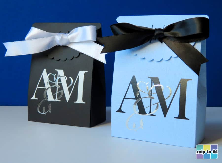 closeup of 2 wedding favor boxes made of blue and black cardstock respectively, each emblazoned with a version of the wedding monogram. The black box features a white monogram with silver script, finished with a white ribbon tied in a bow. The pale blue favor box features a black and silver monogram finished with a black sating bow