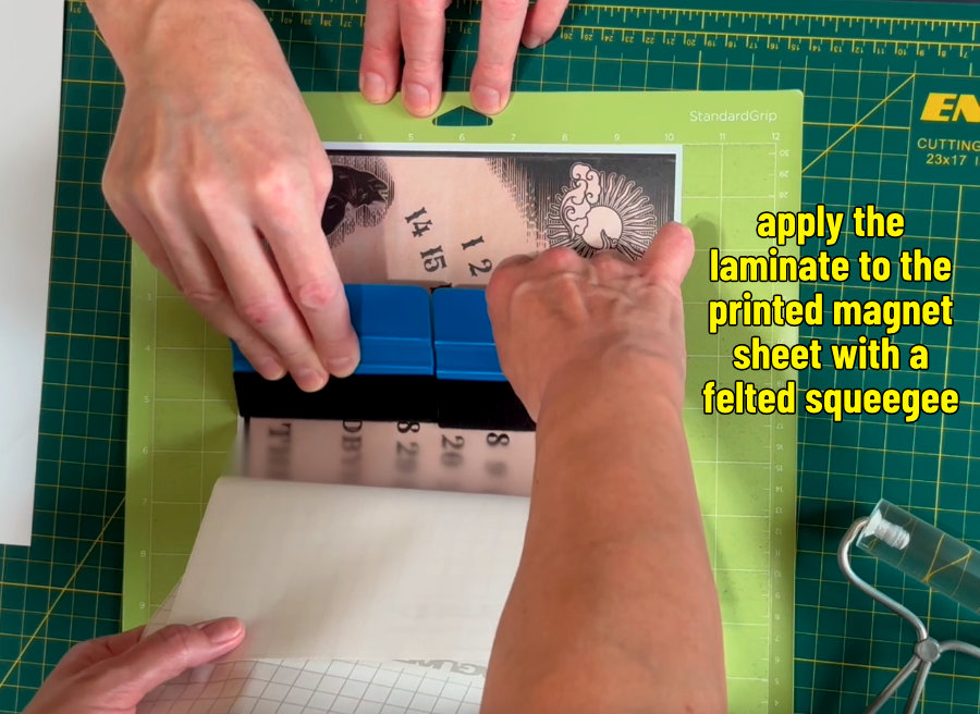photo of 2 people applying a matte Oraguard laminate sheet on top of the printed halloween countdown calendar magnet sheet. Felted squeegees are used to smooth the overlay on top of the printed magnet sheet
