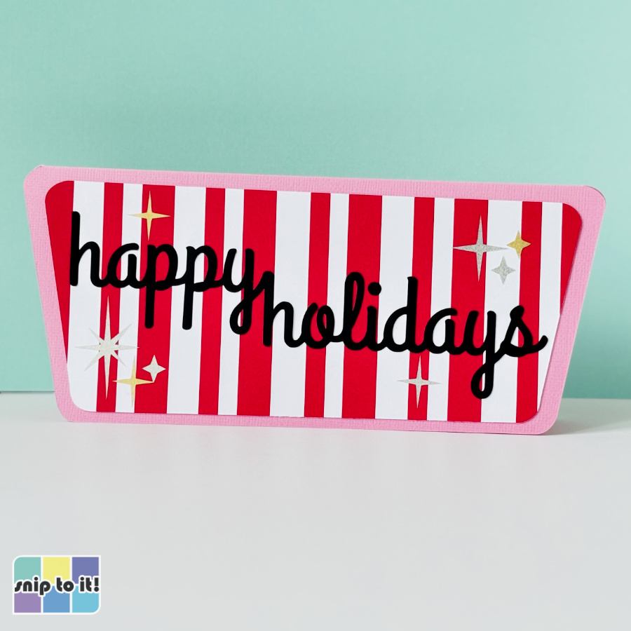 Mid-century–inspired handmade Christmas card featuring a red-and-white vertical candy-stripe inlay, pink rhombus-shaped card base, black “happy holidays” script sentiment, and small retro starburst accents in gold, white, and silver, displayed upright against a mint green background.