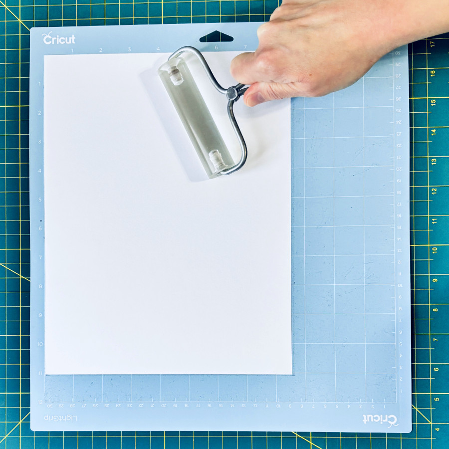 sheet of plain white cardstock being applied to a blue cricut mat with a clear acrylic brayer